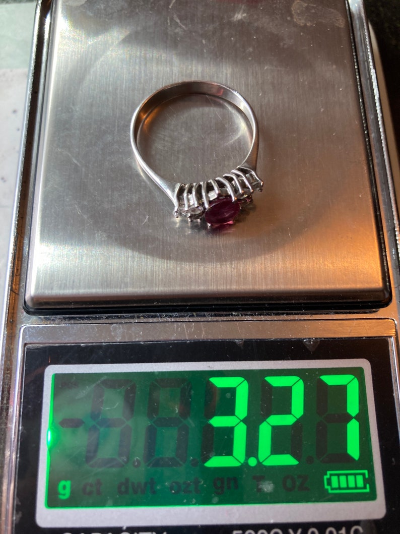 May include: A silver ring with a large, dark red gemstone surrounded by smaller clear gemstones. The ring is on a digital scale that reads 3.22 grams.