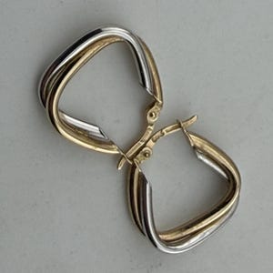 May include: A pair of gold and silver hoop earrings. The earrings have a square shape with a twisted design. The gold and silver colors create a two-tone effect. The earrings are displayed on a light gray surface.