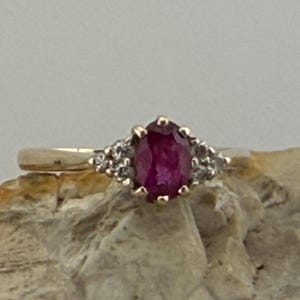 May include: A gold ring with a central oval ruby, complemented by smaller accent stones. The ruby is a rich red hue, and the ring has a timeless design. The ring is displayed on a textured surface. The ring's design is classic.