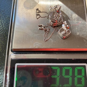 May include: A silver heart-shaped pendant necklace and matching earrings are displayed on a digital scale. The scale reads 3.98, likely indicating the weight in grams. The jewelry is set with small, clear stones.