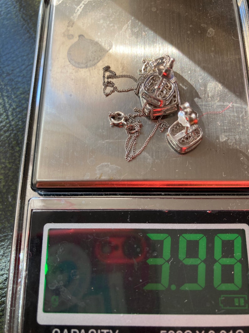 May include: A silver heart-shaped pendant necklace and matching earrings are displayed on a digital scale. The scale reads 3.98, likely indicating the weight in grams. The jewelry is set with small, clear stones.