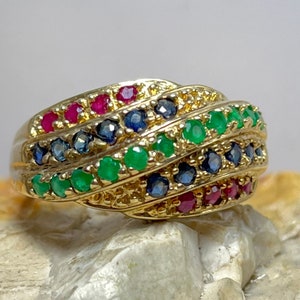 May include: A gold ring with a twisted design, adorned with rows of red, blue, and green gemstones. The ring is displayed on a light-coloured surface, highlighting the colourful gemstones and the polished gold.