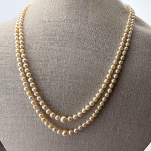 Op de afbeelding: Een dubbele ketting met crèmekleurige imitatieparels van verschillende maten. De ketting wordt getoond op een mannequin, die het elegante ontwerp en de klassieke stijl laat zien. De parels hebben een subtiele glans.