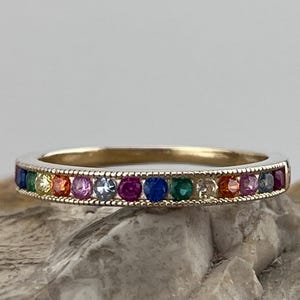 May include: A gold band ring featuring a row of colourful gemstones. The gemstones are set in a milgrain setting and arranged in a rainbow pattern.
