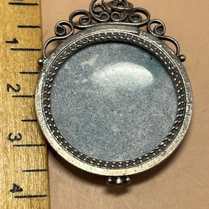 Peut inclure: Un pendentif argent&eacute; avec un cadre rond orn&eacute; et une surface lisse de couleur grise. Le pendentif a une boucle en haut pour attacher une cha&icirc;ne.