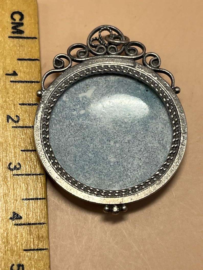 Peut inclure: Un pendentif argent&eacute; avec un cadre rond orn&eacute; et une surface lisse de couleur grise. Le pendentif a une boucle en haut pour attacher une cha&icirc;ne.