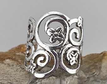 May include: A silver ring with an openwork design featuring floral and swirl patterns. The ring has a wide band and is made of a shiny, metallic material. The intricate details and open spaces create a delicate and elegant look.