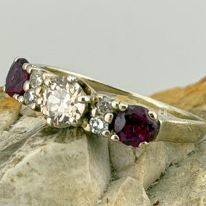 以下が含まれることがあります： 3つの宝石が付いたゴールドリング。リングは、2つの小さな濃い赤色の宝石に挟まれた、大きな透明なセンターストーンが特徴です。追加の小さな透明な石がバンドに沿ってセットされています。