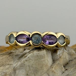 Puede incluir: Un anillo de oro con una serie de piedras preciosas de forma ovalada. El anillo alterna piedras moradas y verde-azuladas, engastadas en una banda de oro. El anillo se muestra sobre una superficie clara y texturizada.