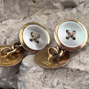 Può includere: Un paio di gemelli d'oro con un design a bottone in madreperla bianca. Ogni bottone ha una croce dorata cucita al centro.