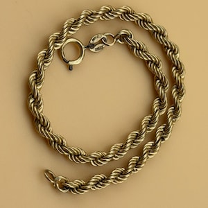 May include: A gold-toned rope chain bracelet featuring a twisted metal link design. The bracelet is secured with a circular spring ring clasp and includes a small metal tag near the closure that displays a stamped hallmark for authenticity.