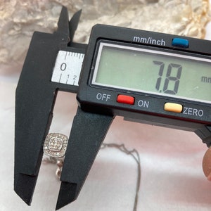 May include: A square-shaped pendant necklace with a cluster of small, clear stones. The pendant is being measured with a digital caliper, showing a measurement of 7.8 mm. The necklace is on a silver chain.