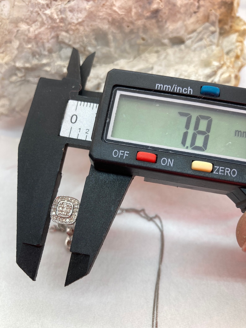May include: A square-shaped pendant necklace with a cluster of small, clear stones. The pendant is being measured with a digital caliper, showing a measurement of 7.8 mm. The necklace is on a silver chain.