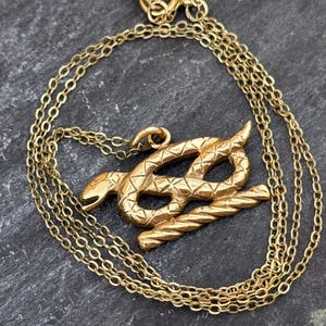 May include: A gold-toned necklace featuring a pendant shaped like a coiled snake resting on a textured, rope-like bar. The snake has a cross-hatched pattern on its body, and the pendant is suspended from a delicate, thin gold-toned chain.