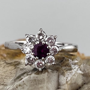 Op de afbeelding: Een zilveren ring met een bloemvormig ontwerp. Het midden van de bloem is een ronde, dieprode edelsteen, omringd door zes heldere, ronde edelstenen. De ring ligt op een getextureerd, lichtbruin oppervlak.