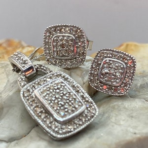 May include: A silver pendant necklace and matching stud earrings set. The jewelry features a square design with a central cluster of small stones surrounded by a border of smaller stones. The pendant is rectangular and the earrings are square.