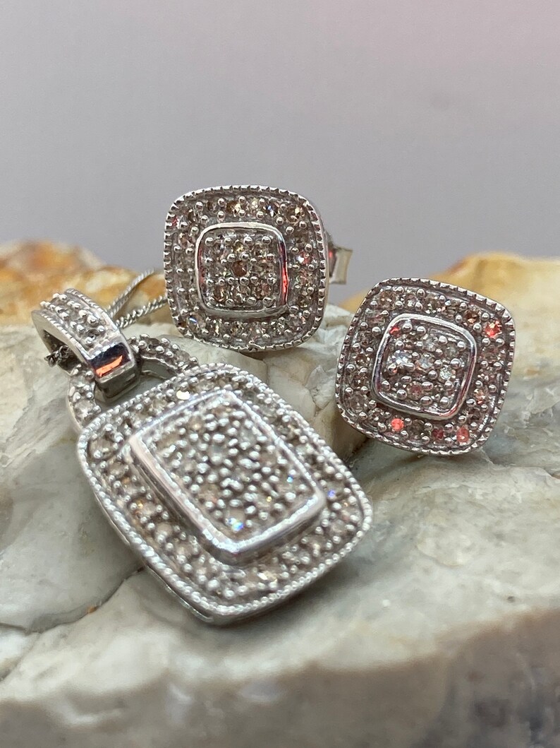 May include: A silver pendant necklace and matching stud earrings set. The jewelry features a square design with a central cluster of small stones surrounded by a border of smaller stones. The pendant is rectangular and the earrings are square.