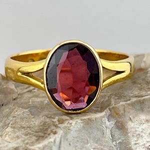 Könnte beinhalten: Ein goldener Ring mit einem tiefvioletten ovalen Edelstein, der in einer Zargenfassung gefasst ist. Der Ring hat eine geteilte Schiene.