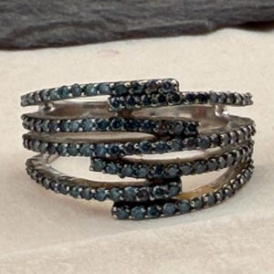 May include: A multi-band ring featuring five overlapping silver-toned bands, each encrusted with small, dark blue stones. The bands are arranged in a staggered, layered design that creates a continuous, textured appearance across the entire surface.