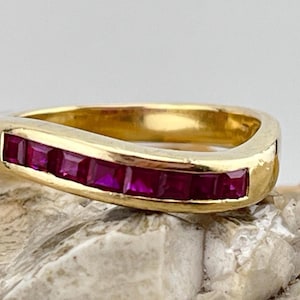 Könnte beinhalten: Ein goldener Ring mit geschwungenem Design, besetzt mit einer Reihe quadratisch geschliffener, tiefroter Edelsteine. Der Ring wird auf einer strukturierten, hellen Oberfläche präsentiert, die das polierte Gold und die lebendige Farbe der Steine hervorhebt.
