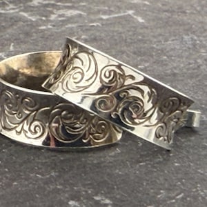 May include: Two silver rings with an ornate, swirling floral pattern. The rings are stacked, with one partially inside the other. The detailed design is carved into the metal, creating a textured surface. The rings are set against a dark, textured background.