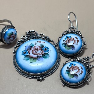 Peut inclure: Un ensemble de bijoux en argent avec &eacute;mail bleu et blanc. L'ensemble comprend une bague, un pendentif et une paire de boucles d'oreilles. Chaque pi&egrave;ce pr&eacute;sente un motif floral peint &agrave; la main avec une rose rose et des fleurs bleues.