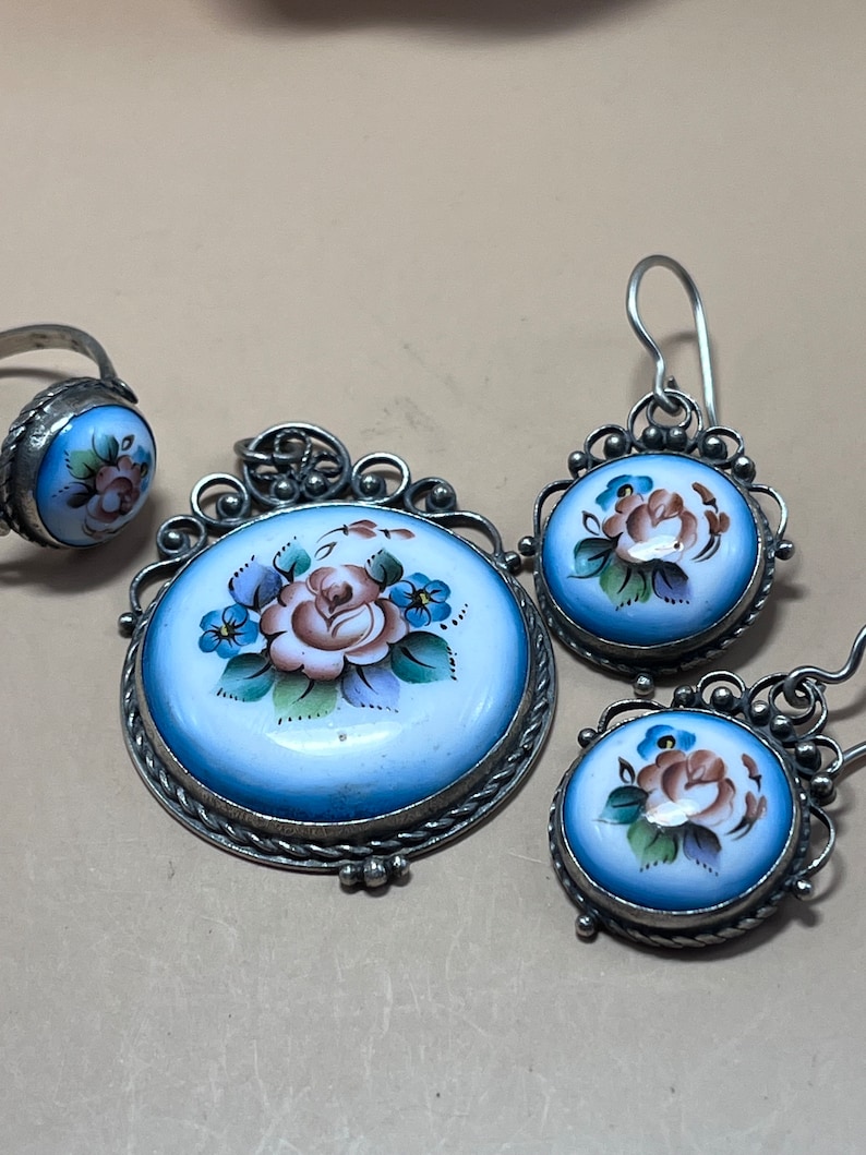 Peut inclure: Un ensemble de bijoux en argent avec &eacute;mail bleu et blanc. L'ensemble comprend une bague, un pendentif et une paire de boucles d'oreilles. Chaque pi&egrave;ce pr&eacute;sente un motif floral peint &agrave; la main avec une rose rose et des fleurs bleues.