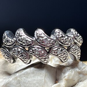 Puede incluir: Un anillo plateado con un patrón repetitivo de elementos en forma de frijol. Cada elemento tiene una superficie texturizada y una pequeña piedra transparente. El anillo se muestra sobre una superficie clara y texturizada sobre un fondo oscuro.