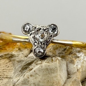 May include: A vintage ring with a gold band and a silver setting. The silver setting has a triangular design with three clear gemstones and smaller accent stones. The ring is displayed on a textured, light-coloured surface.