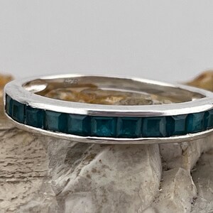 May include: A silver ring featuring a row of square, teal gemstones. The ring is displayed against a neutral backdrop, highlighting the gemstones' colour and the ring's design. The ring is a piece of jewellery.
