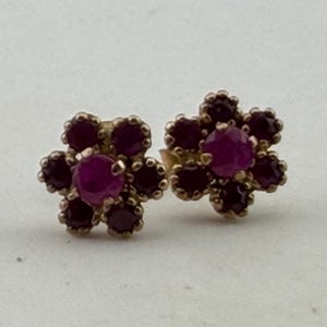 Op de afbeelding: Een paar bloemvormige oorstekers. Elke oorbel heeft een centrale, ronde, dieproze edelsteen omringd door kleinere, donkerrode edelstenen. De oorbellen hebben een goudkleurige zetting en worden tegen een lichte achtergrond weergegeven.