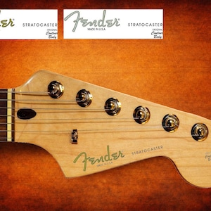 Può includere: Primo piano della paletta di una chitarra Fender Stratocaster. La paletta è in legno chiaro, con meccaniche dorate e corde. Il logo Fender e "Stratocaster" sono visibili in verde. Lo sfondo è una sfumatura di marrone e arancione.