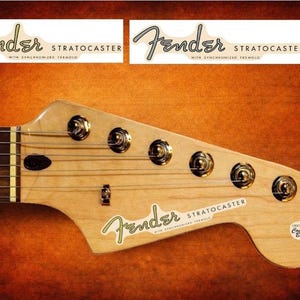 Può includere: Primo piano della paletta di una chitarra Fender Stratocaster. La paletta è in legno chiaro, con sei meccaniche e il logo Fender. È visibile anche il testo "Stratocaster".