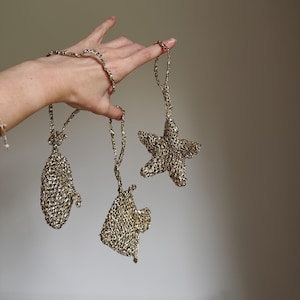 May include: A set of three hanging ornaments crafted from metallic gold yarn, featuring a mitten, a star, and a small house shape. Each piece is connected by a matching gold cord, showcasing a textured, open-knit construction with a shimmering finish.