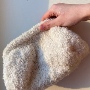 May include: A cream-coloured, textured clutch bag. The bag has a soft, fluffy appearance. The material appears to be a knit or woven fabric. The clutch is held by a hand, suggesting its size and portability. The neutral colour makes it versatile.