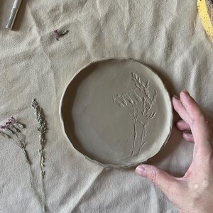 May include: A round, light brown clay plate featuring an embossed botanical design of two stems with leaves and flowers. The plate has a slightly uneven, hand-formed edge and a smooth, matte surface texture, showcasing a natural, earthy appearance.