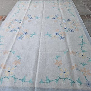 May include: A large, rectangular, off-white rug with a floral pattern. The design features peach, blue, and teal flowers and vines. The rug has a light blue border and is likely made of a soft, woven material.