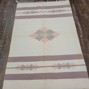 May include: A white rug with a geometric pattern in shades of pink, brown, and gray. The rug has a fringe border.