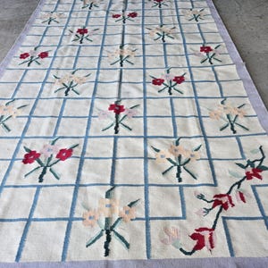 May include: A rectangular rug with a cream-colored background and a grid pattern of light blue lines. The grid is filled with embroidered floral designs in shades of red, pink, and cream. The rug has a lavender border.