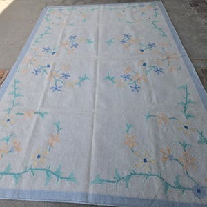 May include: A rectangular, off-white rug with a light blue border. The rug features a floral pattern in shades of blue, peach, and green. The floral design is arranged around the edges and in the center of the rug.