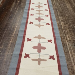 May include: A long, rectangular rug with a white background and a geometric pattern in shades of red, pink, and gray. The rug has a blue border.