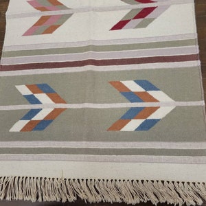 May include: A white woven rug with a fringe border. The rug has a geometric pattern of arrows in shades of blue, red, orange, and white on a gray background.