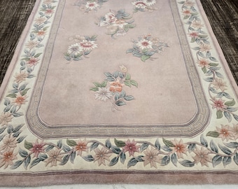 May include: A pale pink rug with a floral design. The rug has a border of leafy vines with flowers in shades of pink, white, and green. The center of the rug features a repeating pattern of stylized flowers.