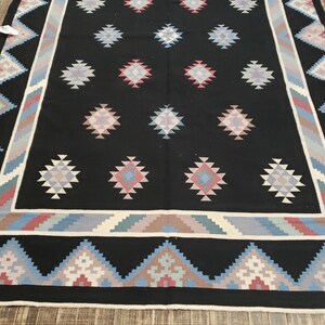 May include: A black area rug with a geometric pattern. The rug features diamond-shaped designs in shades of pink, blue, and gray, set against a black background. The border has a repeating triangle pattern in similar colors.