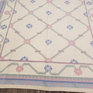 May include: A white rug with a geometric pattern of pink, blue, and green flowers. The rug has a border of blue, pink, and green.