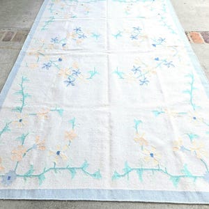 May include: A rectangular white rug with a light blue border. The rug features floral embroidery in shades of peach, blue, and teal. The floral pattern is concentrated in the corners and along the edges of the rug.