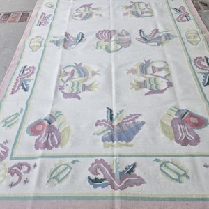 May include: A rectangular rug with a white background and a repeating pattern of colorful fish, shells, and floral designs. The border is a light pink with a teal and white trim. The rug is made of a soft material.