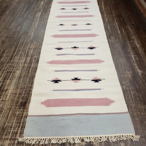 May include: A white runner rug with a geometric pattern in shades of pink, blue, and black. The rug has a fringe border.