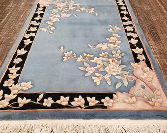 Op de afbeelding: Een lichtblauw tapijt met een bloemenpatroon met delicate roze en witte bloemen en een zwarte rand. Het tapijt heeft een franje rand.