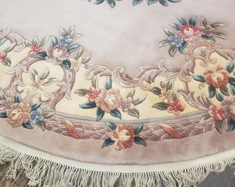 May include: A round rug with a light pink background and a cream-colored border. The rug features a floral design with pink, blue, and yellow flowers. The rug has a fringe border.